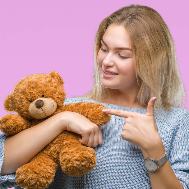 Young caucasian woman holding cute teddy bear over isolated background very happy pointing with hand and finger - 写真・画像