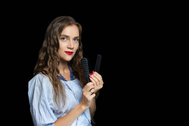 Beautiful girl with very confident face holding black comb, hairbrushes. On her eyes arrows, nude skin beige concealer, bright red lips. Profession Hairdresser is ready to work in a new saloon studio - Foto, afbeelding