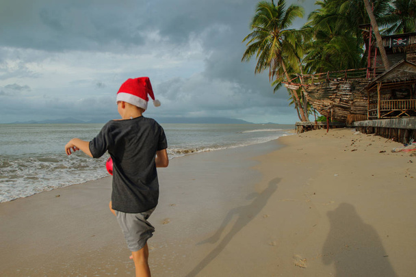 Criança feliz no chapéu do Papai Noel na praia vazia Ban Tai em Koh Samui, Tailândia desfrutando de primeiros raios de luz solar após a tempestade tropical Pabuk
 - Foto, Imagem