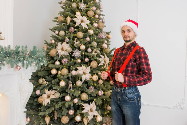 joven hombre guapo con un sombrero rojo de Año Nuevo en pantalones de mezclilla con una camisa blanca y enaguas se sienta en el árbol de Navidad con regalos de humor festivo
 - Foto, imagen