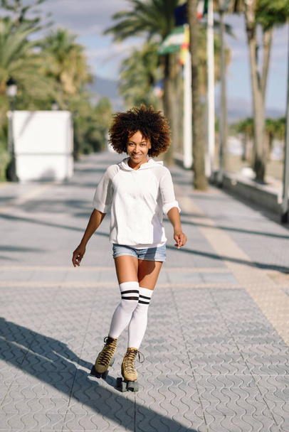 Smiling black woman on roller skates riding outdoors on beach promenade with palm trees. Smiling girl with afro hairstyle rollerblading on sunny day. - 写真・画像
