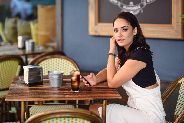 Kaunis tyttö sinisilmäinen istuu kaupunkien kahvila älypuhelimella. Nuori nainen ruskea aaltoileva kampaus yllään valkoinen denim mekko. Elämäntapakäsite
. - Valokuva, kuva