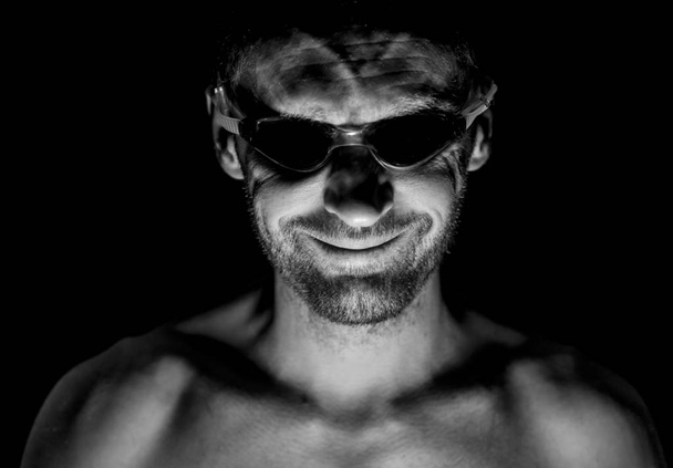Portrait of unshaven adult caucasian man in swimming glasses. He smiles like maniac and seems like madness or crazy. Black and white shot, low-key lighting. Isolated on black. - Foto, afbeelding