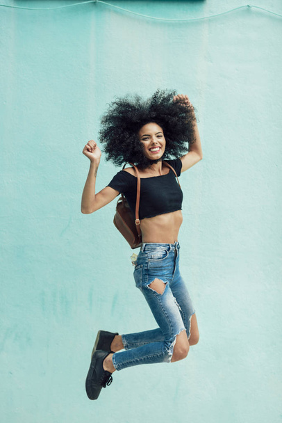 Young mixed woman with afro hair jumping outdoors. Female wearing casual clothes in urban background. Lifestyle concept - 写真・画像