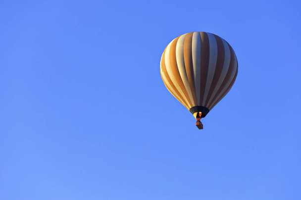 Пламя огня нагревает воздух в пестром желто-белом красивом воздушном шаре и поднимает корзину туристов в голубое небо
. - Фото, изображение