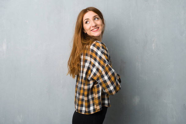 Young redhead girl over grunge wall looking over the shoulder with a smile - Фото, зображення