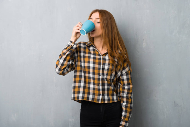 Молода руда дівчина над гранжевою стіною тримає гарячу чашку кави
 - Фото, зображення