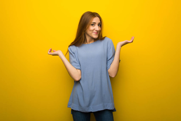 Young redhead girl over yellow wall background having doubts while raising hands and shoulders - Photo, Image