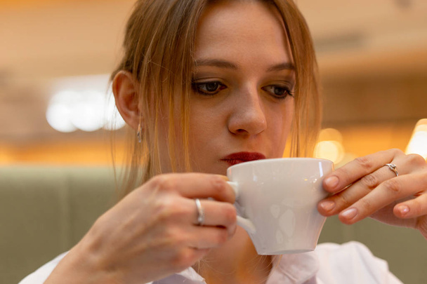 Girl office manager in a business blouse thoughtfully drinks a cappuccino during a coffee break in the office. Emotion is sadness. - Zdjęcie, obraz