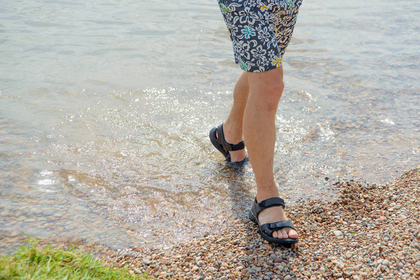 Feet in sandals on the beach. Stony shore of the lake. Man in shorts. Beach colored shorts on a man. Wet sandals. The opening of the beach season. Walk on the rocky beach. - Foto, Imagen