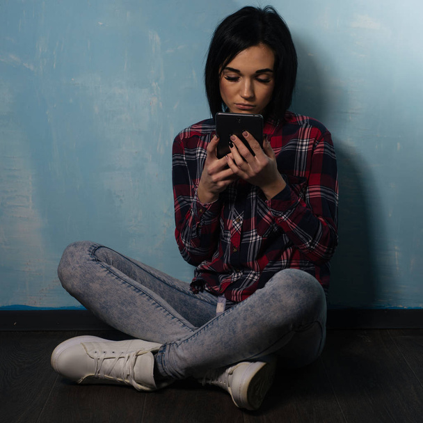 Smutna dziewczyna cierpi na uzależnienie od sieci społecznościowych, siedząc na podłodze ze smartfonem - Zdjęcie, obraz