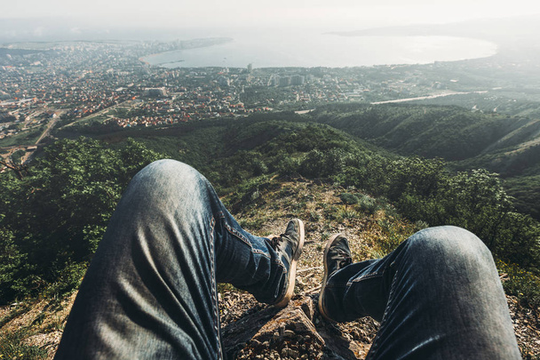 Man traveler sits on top and enjoys view of the picturesque landscape and the city. Point of view shot. Travel, adventure, tourism concept - Foto, Bild