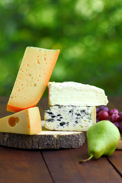 Camembert, brie, hard cheese and blue cheese on a wooden board. Slices of various cheeses and grapes on a blurred background. Dorblu and soft cheese on wooden boards. Cheese and grapes on bokeh - Foto, imagen