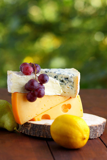 Camembert, brie, hard cheese and grapes on a wooden stand. Pieces of different cheeses, pear and lemon on a blurred background. Dorblu and soft cheese on wooden boards. Cheese and fruit on bokeh - Foto, Imagem