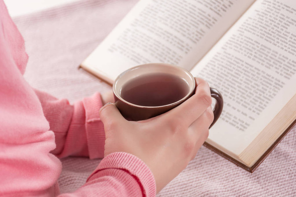 Young woman holding cup of tea and reading book at home. Girl wears pink woolen gently sweater. Education and cold weather concept. Close up, selective focus - Foto, Imagen