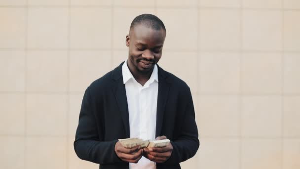 Portrait of african american businessman in suit counting money standing near office centre. He celebrating his successful win with a lot of dollars and looking into the camera. - Кадры, видео