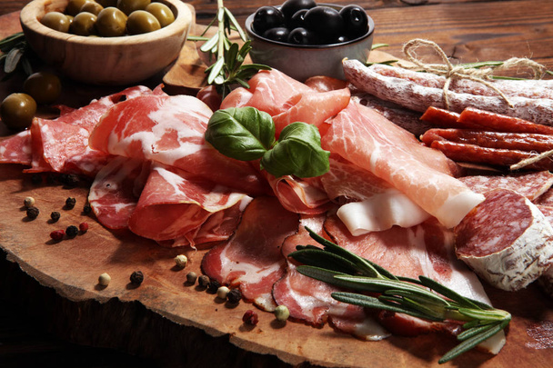 Marble cutting board with prosciutto, bacon, salami and sausages on wooden background. Meat platter appetizers and olives - Foto, immagini