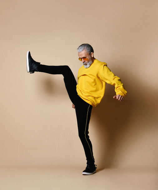 senior millionaire man in yellow cloth and aviator stylish sunglasses exercise stretching hit punch with leg - 写真・画像