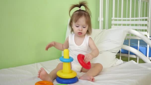Baby sitzt auf dem Krankenhausbett und spielt mit Pyramide. Krankes Mädchen erholt sich unter ärztlicher Aufsicht im Krankenhaus - Filmmaterial, Video