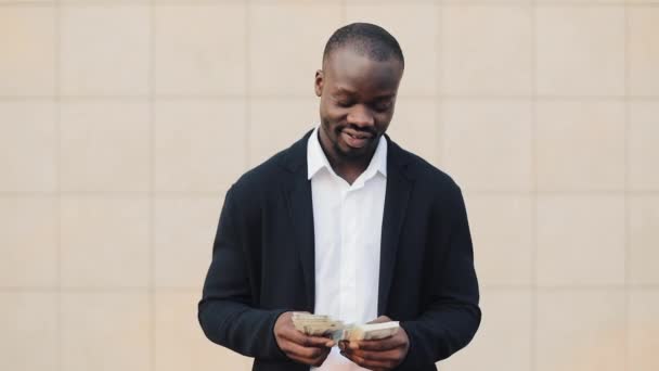 Portrait of african american businessman in suit counting money standing near office centre. He celebrating his successful win with a lot of dollars and looking into the camera - Filmagem, Vídeo
