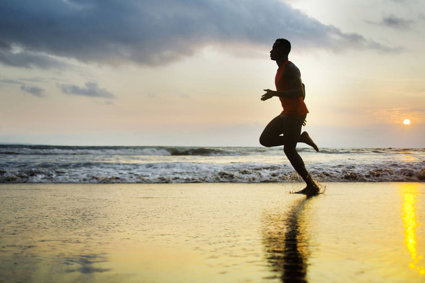 silhouette of young attractive fit athletic and strong black afro American man running at sunset beach training hard and sprinting on sea water in professional athlete and runner workout  - 写真・画像