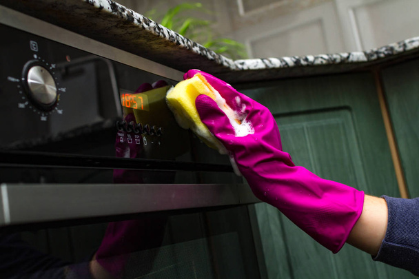 Háziasszony kéz a gumikesztyűt mosás elektromos sütővel, mosó-és tisztítószerek és a konyhai szivacs  - Fotó, kép
