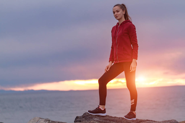 Fitness attractive woman in sneakers climbing on a rock after a workout and looking into the distance on a sea background at sunset. Sports and active lifestyle - 写真・画像