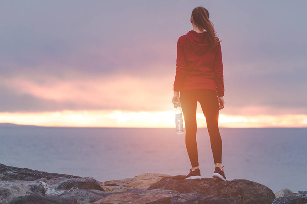 Fitness-Frau in Turnschuhen steht auf einem Stein, hält eine Wasserflasche in der Hand und ruht sich nach dem Training auf einem Meeresgrund bei Sonnenuntergang aus. Sport und aktiver Lebensstil. - Foto, Bild