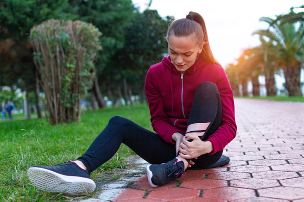 A young, fitness girl in sneakers and sportswear was injured while jogging and sports training. Leg injury and muscle strain, and ankle sprain accident during sports exercise.  - Фото, зображення