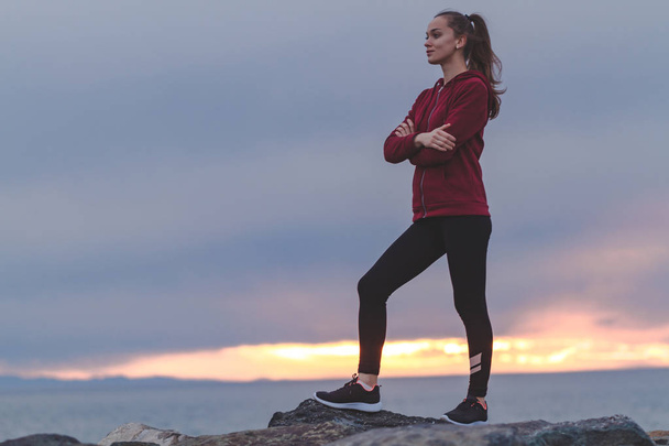 Fitness attractive woman in sneakers climbing on a rock after a workout and looking into the distance on a sea background at sunset. Sports and active lifestyle - Photo, Image