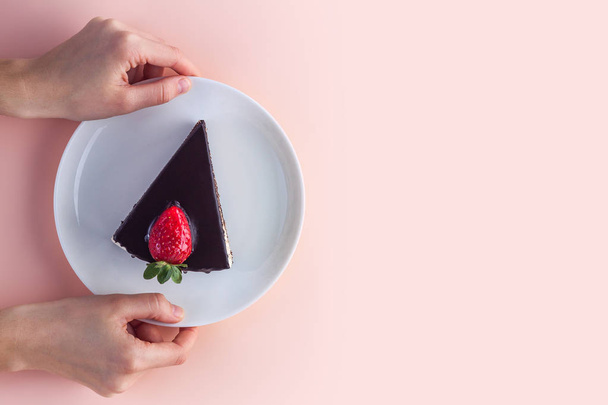 A slice of sweet strawberry cake with dripping chocolate glaze in a white plate in hands on a pink background. Sweet dessert. Copy space  - Fotografie, Obrázek
