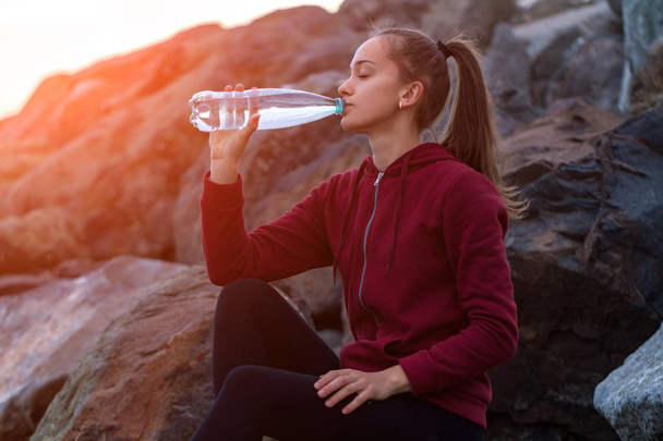 Фитнес привлекательная женщина в кроссовках сидит на камне, пьет воду и отдыхает после тренировки на закате. Спорт и активный образ жизни
 - Фото, изображение