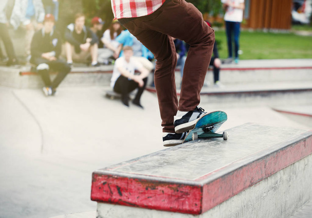 Skateboarder boy grinding on a ledge in outdoor skate park. Crowd on background, sport competition for youth. Popular extreme sport, dangerous trick. Focus on skateboard - 写真・画像