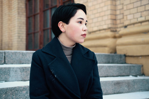 Melancholic portrait of young asian woman with short hair style sitting on the stairway. Sensual millennial hipster girl wearing a blue coat. - Foto, immagini