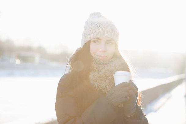 Cute brunette chick enjoying hot drink.White paper takeaway cup from local barista.Place your cafe logo on clean mug for refreshing drinks.Lens flare and bright sunlight.Model in warm winter wear - Фото, зображення