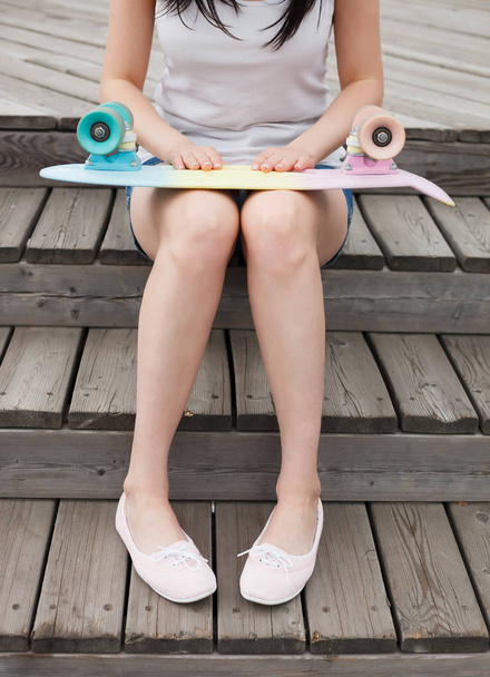 Jonge vrouw zitten buiten houden van korte cruiser skateboard op zomerdag. Populaire kleine skate deck, trendy outfit, focus op de voeten van onherkenbaar model - Foto, afbeelding