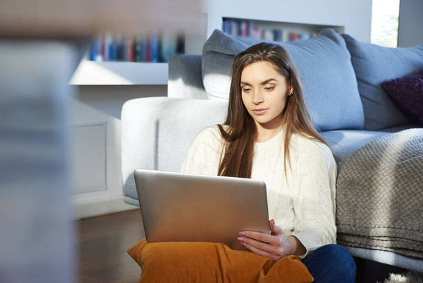 Portrait of thinking young woman using her laptop while sitting at sofa at home. Home office. - Fotoğraf, Görsel