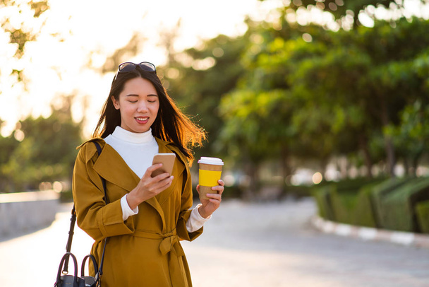Cheerful girl using phone and having a cup of coffee outdoors - Photo, Image