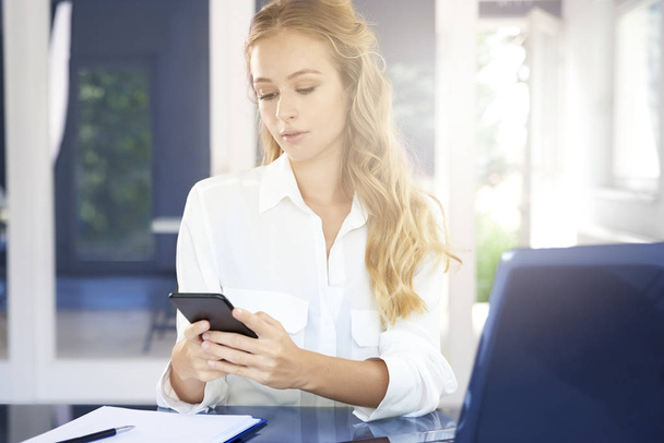 Portret van mooie jonge zakenvrouw achter bureau en SMS-berichten tijdens het werken op de laptop. - Foto, afbeelding