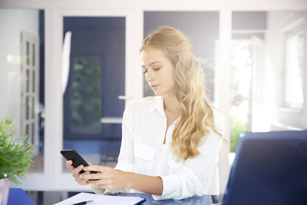 Portrait of beautiful young businesswoman sitting at office desk and text messaging while working on laptop. - Foto, Bild