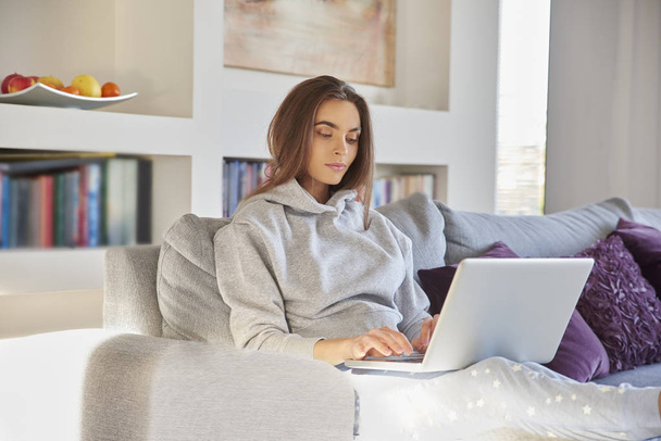 Shot of a casual young woman wearing hoodie and pajama pants while relaxing on sofa and using her laptop. - Foto, Imagem