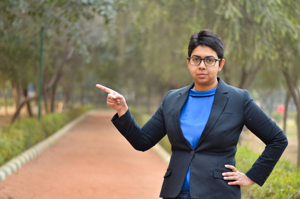 Retrato de uma jovem indiana confiante Mulher profissional corporativa com cabelo curto apontando para a direita com o dedo em um ambiente ao ar livre vestindo um negócio preto / terno formal
 - Foto, Imagem