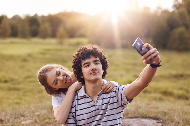 Couple of teenagers have fun together, make selfie, holds smart phone, show tongue, dressed in casual outfit, pose against field background with sunshine. Curly guy uses cellular for taking photo - Photo, image