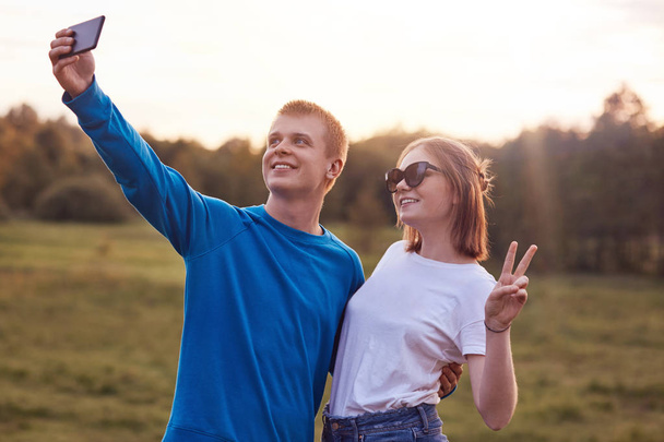 Outdoor shot of funny best friends make peace gesture, stand closely to each other, take selfie with cell phone, wear sunglasses, spend summer vacation in open air. Teenagers and fun concept - Fotografie, Obrázek