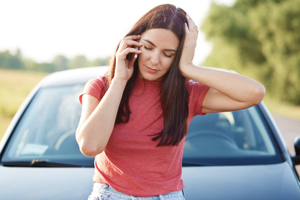 Tired European woman with long hair has telephone conversation via mobile phone, feels tired after repairing car, asks for help, dressed in casual t shirt. People, conversation, transport concept - Photo, Image