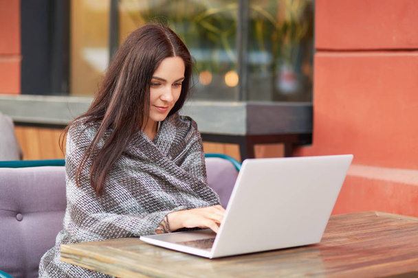 Pretty lady sits in front of opened laptop computer, writes email, uses high speed internet, wrapped in plaid, poses in outdoor cafeteria, works or studies online. People and technology concept - Foto, imagen