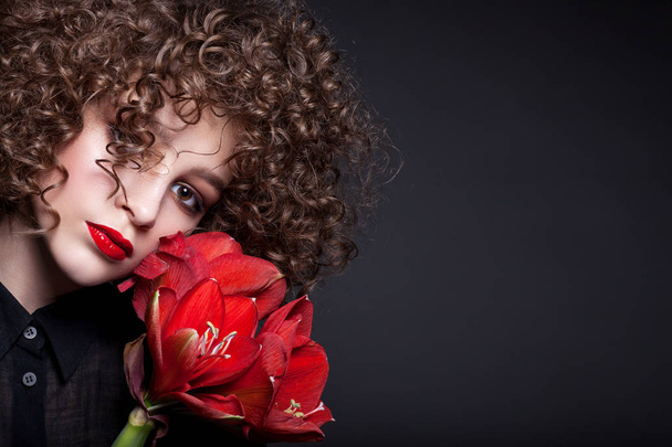 Portrait d'une belle adolescente avec un maquillage lumineux. Avec des boucles afro et rouge à lèvres rouge et une fleur rouge sur le visage. Sur fond noir
. - Photo, image