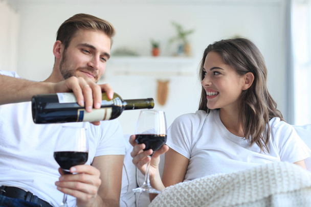 Young loving couple drinking a glass of red wine in their living room - Фото, изображение