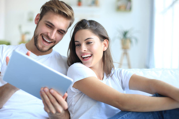 Mladý pár sledovat mediální obsah on-line v tabletu sedí na pohovce v obývacím pokoji. - Fotografie, Obrázek