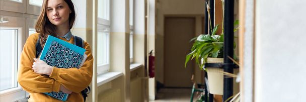 Jovem e atraente estudante do ensino médio em pé à janela no corredor em sua escola sozinho. Conceito de educação
. - Foto, Imagem